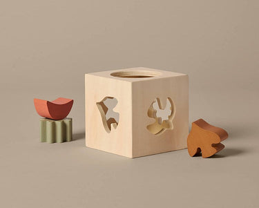 A Ferm Living wooden shape-sorting cube toy with four geometric cutouts, shown on a warm beige background with small matching wooden pieces.