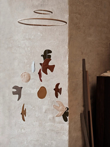 A Ferm Living mobile wall hanging with organic bird-like shapes and circular elements in earthy brown, beige, and black tones, suspended in a minimalist interior setting.