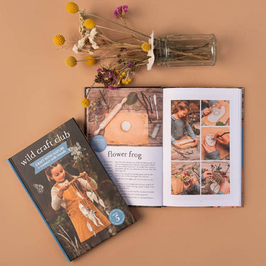 A book and an open book called Wild Craft Club, and some dried flowers ina glass vase on a beige background 
