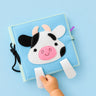 A baby sensory book with a cow face design, held by a child's hand against a solid light blue background.