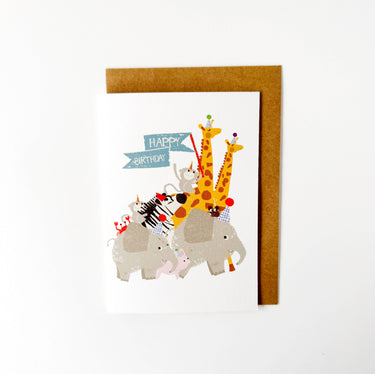 A children's birthday card featuring a colorful illustration of safari animals including giraffes, an elephant, a zebra, and a pink pig, with a blue 'Happy Birthday' banner. The card is shown standing upright with a brown kraft envelope behind it on a white background.