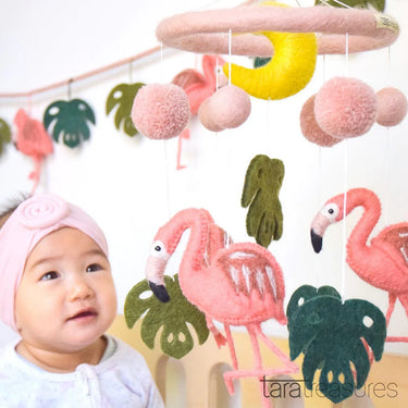 Baby mobile with flamingos, leaves, and pom-poms hanging above a baby