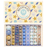An open gift box filled with assorted decorative glass marbles, shown with the lid visible above. The lid is cream-colored with blue, yellow, and white seashell and starfish illustrations and a central circular label reading 'BILLES & CO LES COLLECTIONS 58 PCS OCEAN COQUILLAGES.
