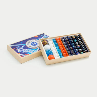 A children's marble game set by Bill & Co, shown with its illustrated cardboard lid leaning against an open box containing multiple rows of colorful glass marbles in white, blue, orange, and black.