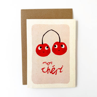 A rectangular greeting card with a kraft brown envelope behind it, featuring an illustration of two smiling red cherries with black stems and the handwritten red text 'mon chéri' on a cream background.