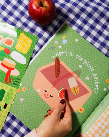 Children's book titled 'What's in the Picnic Basket?' on a checkered tablecloth with an apple beside it.