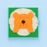 A square fabric sensory book featuring a lion face design, with orange felt ears and hands covering a brown nose, set against a green background with a white faux-fur texture, on a light blue background.