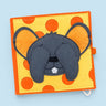 A square fabric sensory activity book featuring a gray mouse face on a yellow background with orange polka dots, photographed on a light blue background.