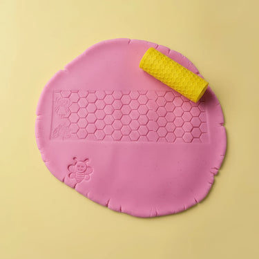 Pink play dough with embossed honeycomb pattern and bee motifs, shown with a yellow honeycomb-textured roller on a pale yellow background.