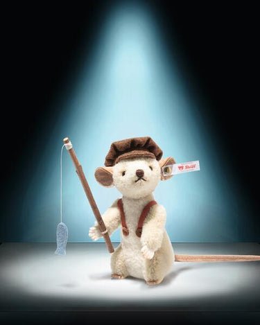 A plush mouse toy dressed as a Huckleberry Finn, holding a fishing rod with a blue fish-shaped lure. The toy has a cream-colored body, a brown hat and suspenders, and a small red-and-white Steiff tag on its ear, set against a dark background with a spotlight effect.