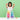 A smiling girl in a white dress stands behind a colorful marble run toy set on a white table against a light green background, holding a pink ball in one hand and a green ball in the other.