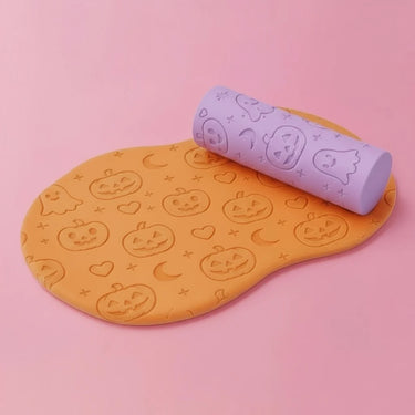 An orange silicone mat with an embossed Halloween pattern of jack-o'-lanterns, ghosts, hearts, and moons, shown with a lavender purple cylindrical foam roller on a solid pink background.