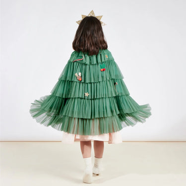 A child viewed from behind wearing a green tiered tulle cape with embroidered decorative details, a pale pink dress, white socks, and a gold star headpiece against a plain light background.